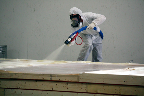 Pracownik w kombinezonie ochronnym i masce natryskuje piankę izolacyjną na drewnianą konstrukcję, widoczny pistolet natryskowy z niebieskim wężem i czerwonym przewodem.